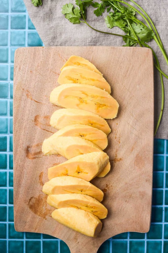 Plantain sliced into thin diagonal pieces on a cutting board. 