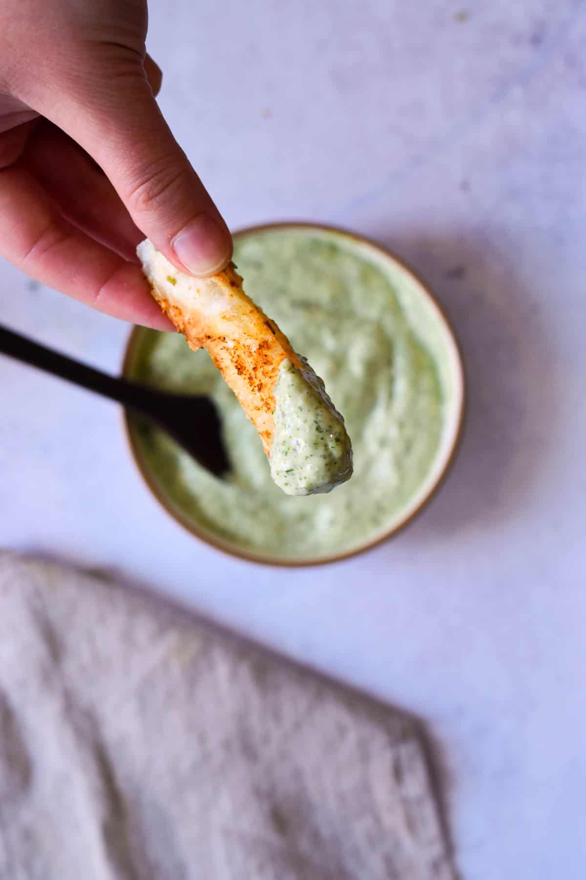 Hand holding a yuca fry that has been dipping in cilantro jalapeno sauce. 