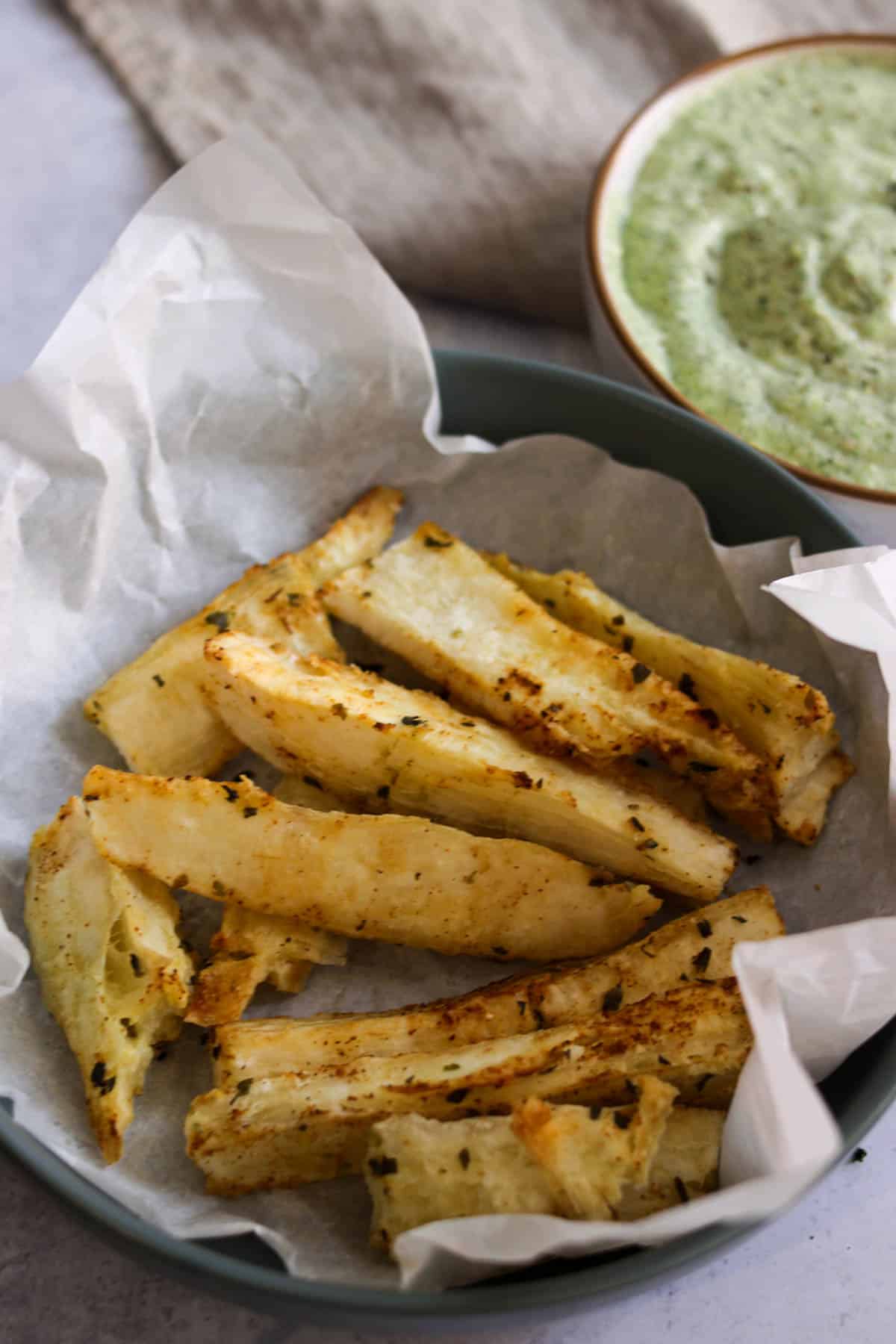 Plate of yuca fries with a lining of parchment paper and green sauce in the background. 